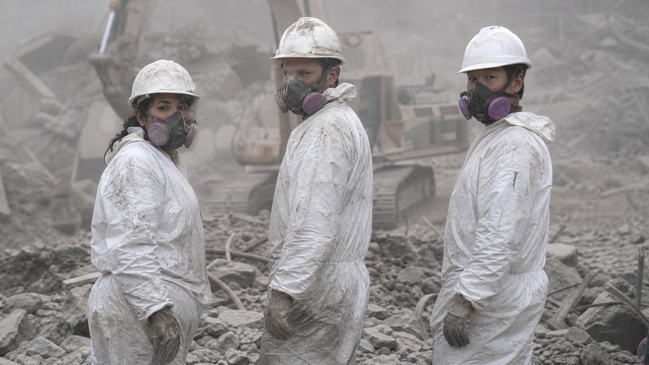 Recovery workers in respirators and dust-covered protective gear standing in a debris field
