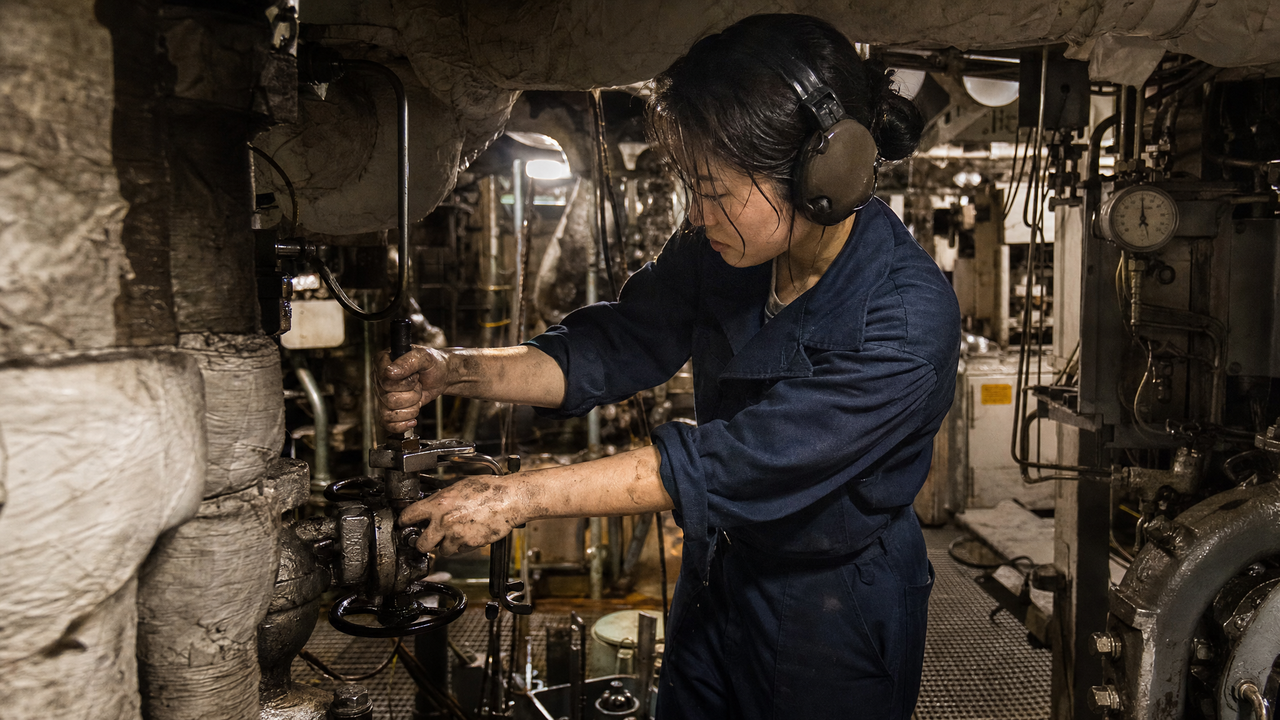 A sailor in coveralls and hearing protection working a valve in a warship engine room surrounded by insulation-wrapped steam pipes