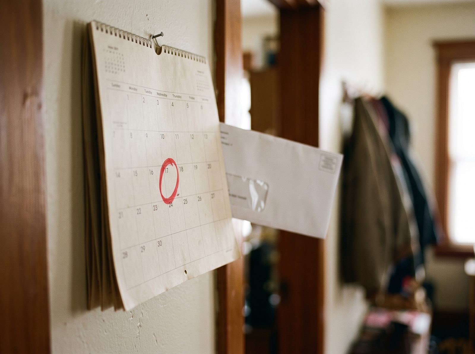A paper wall calendar in a home hallway with one date circled in red marker and a plain white envelope tucked behind it